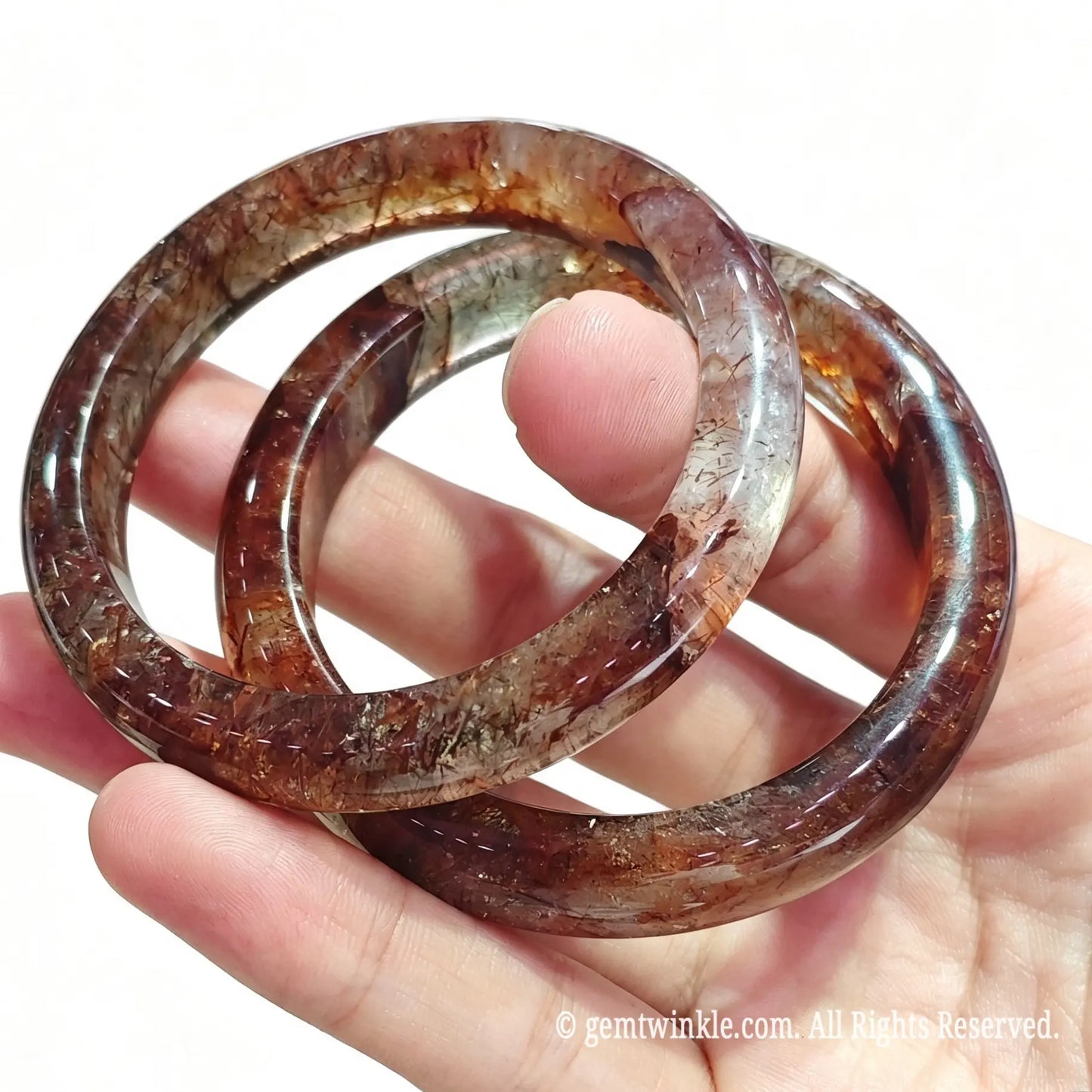 Close up view of two solid super seven quartz bangles held between fingers showing high gloss polish and dense mineral needles