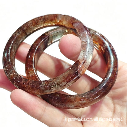 Close up view of two solid super seven quartz bangles held between fingers showing high gloss polish and dense mineral needles