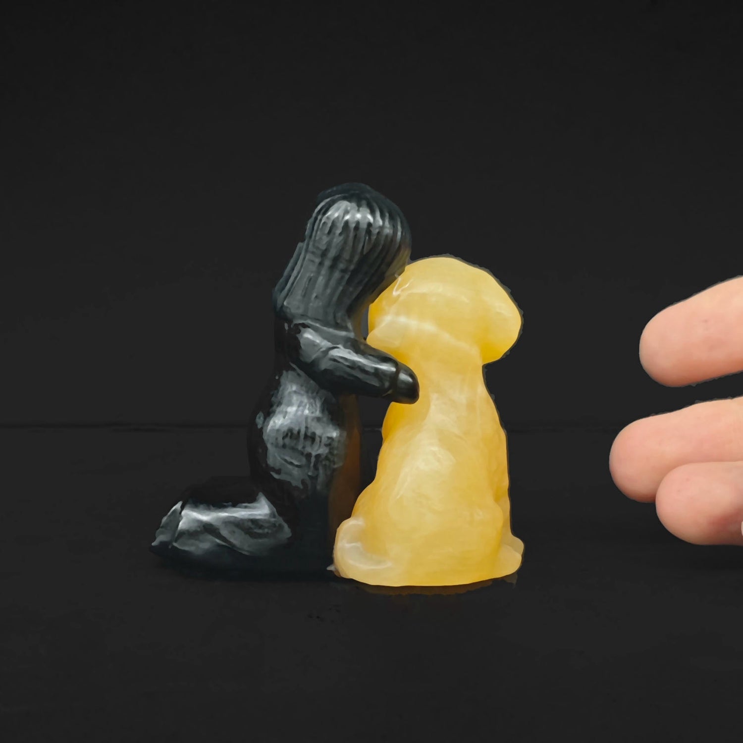 Side view of a 7cm gemstone statue showing a black obsidian girl hugging a honey yellow calcite dog on a plain black background