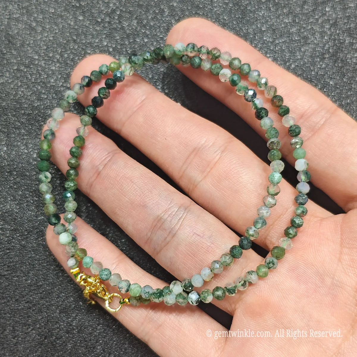 A beaded necklace made of faceted green and white gemstone beads with a gold clasp, shown resting on a person's hand against a dark textured background.