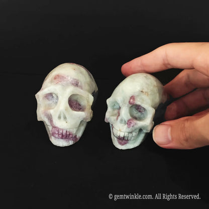 A hand adjusting two 6cm pink tourmaline in quartz human skulls to inspect their anatomical features
