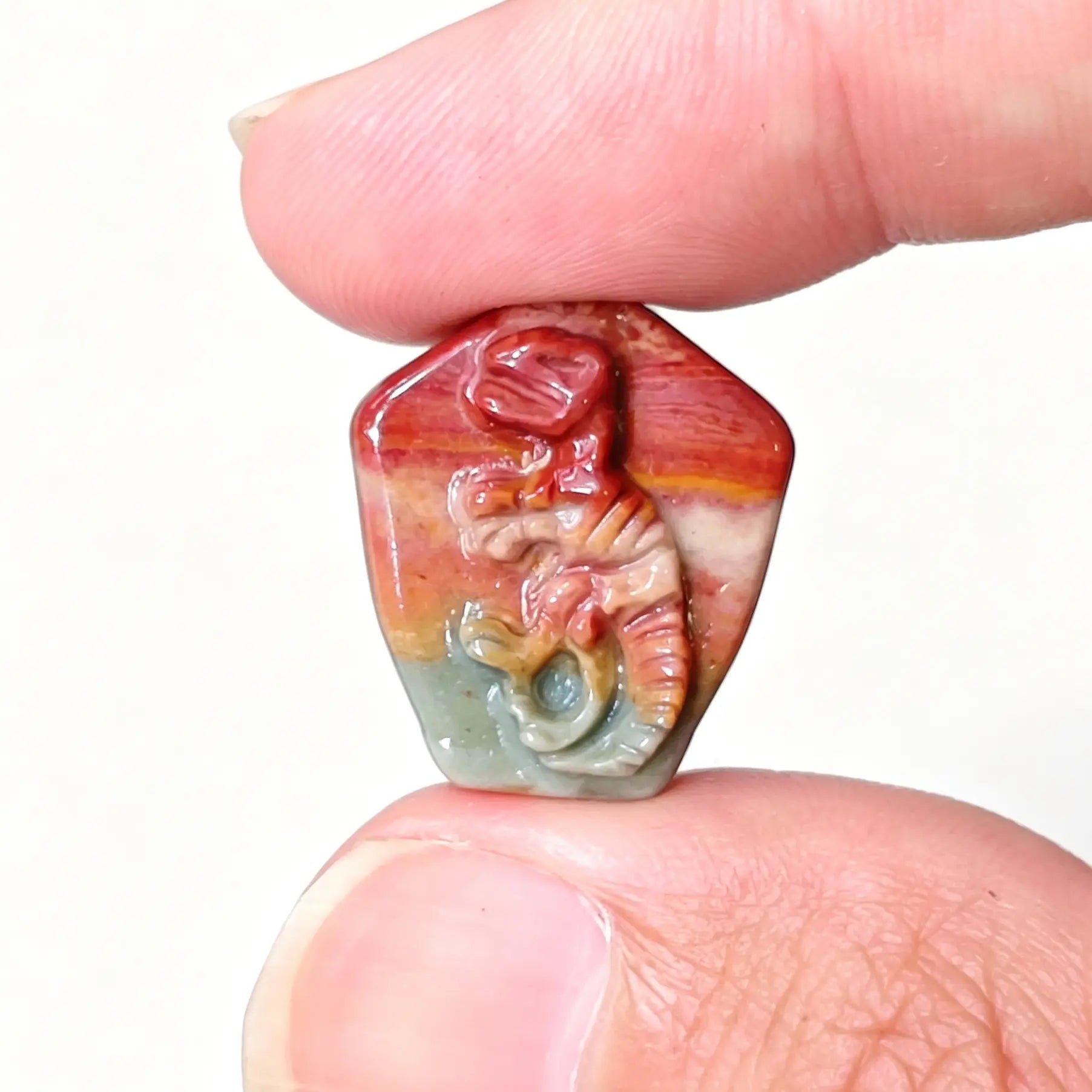 Fingers holding a hand-carved Alashan Gobi agate T-Rex dinosaur fossil skeleton relief displaying red and green earth bands