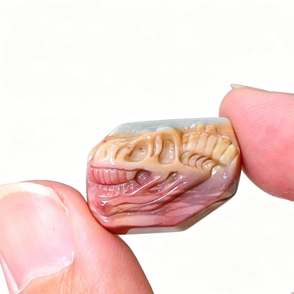 Fingers holding a hand-carved Alashan Gobi agate T-Rex dinosaur skull fossil relief stone showing coral pink and beige bands