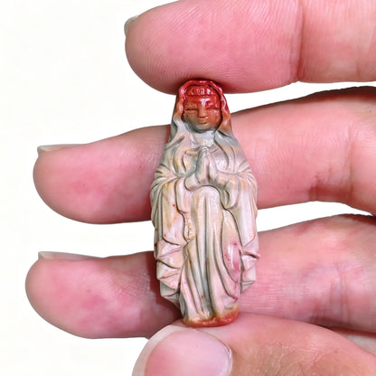 Fingers holding a hand-carved natural Alashan Gobi agate Virgin Mary figurine showcasing red and grey earth tones