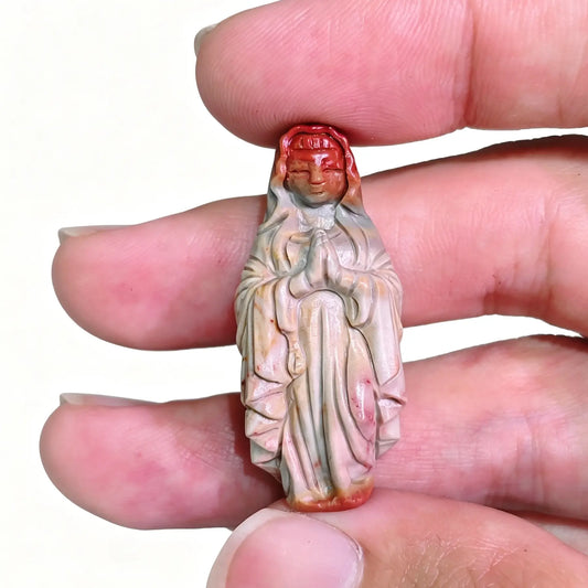 Fingers holding a hand-carved natural Alashan Gobi agate Virgin Mary figurine showcasing red and grey earth tones