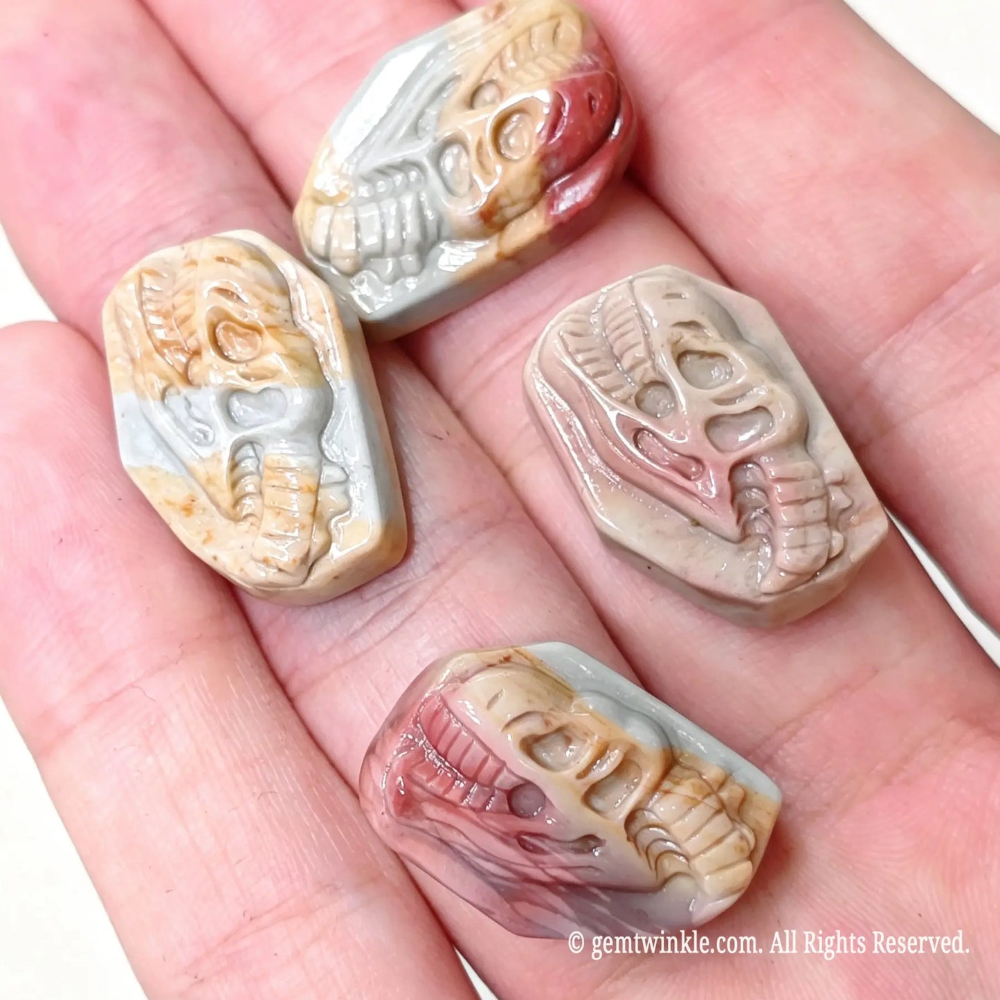 Hand holding four hand-carved Alashan agate dinosaur skull relief cabochons showcasing detailed tooth and bone carvings
