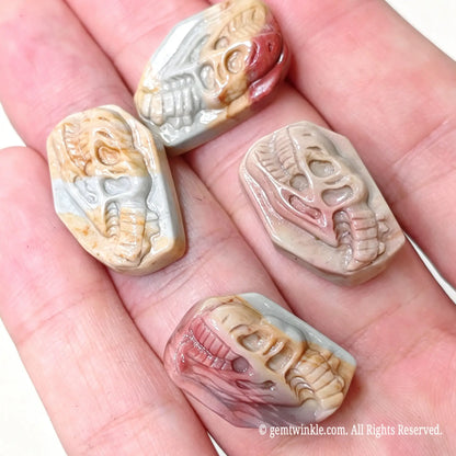 Hand holding four hand-carved Alashan agate dinosaur skull relief cabochons showcasing detailed tooth and bone carvings