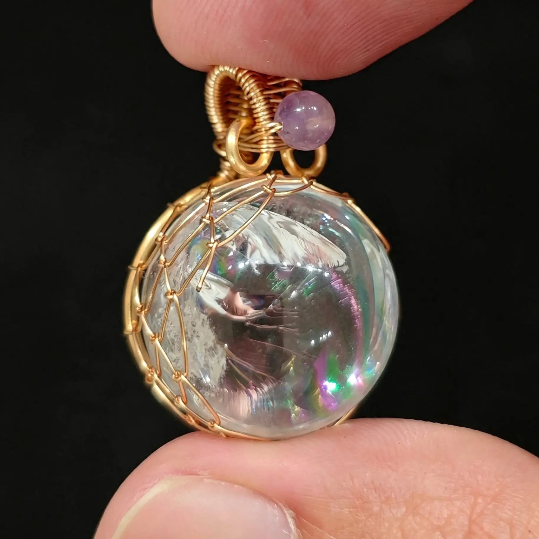 Fingers holding a 20mm clear quartz sphere pendant with vivid rainbow inclusions in gold wire cage wrapping