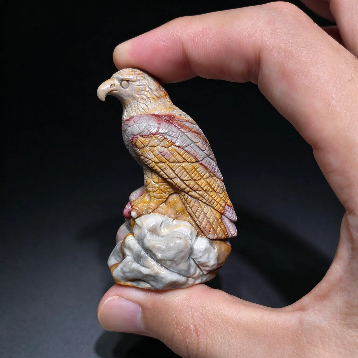 Profile view of a carved eagle statue with golden yellow and rusty red feather detailing held between fingers under studio light