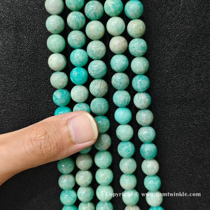 Finger holding natural Grade C amazonite round bead strands demonstrating size and surface polish