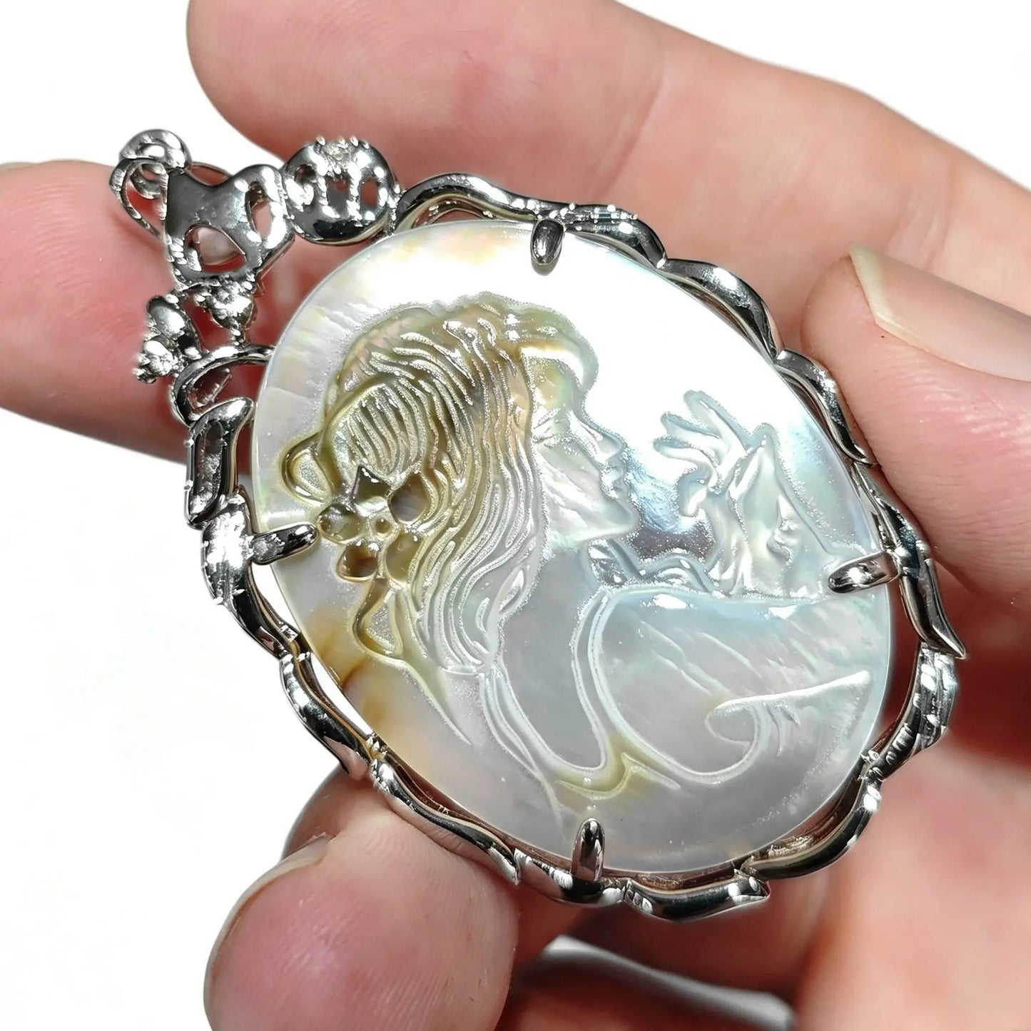 Close up view of carved mother of pearl cameo pendant held between fingers displaying ornate silver tone prongs