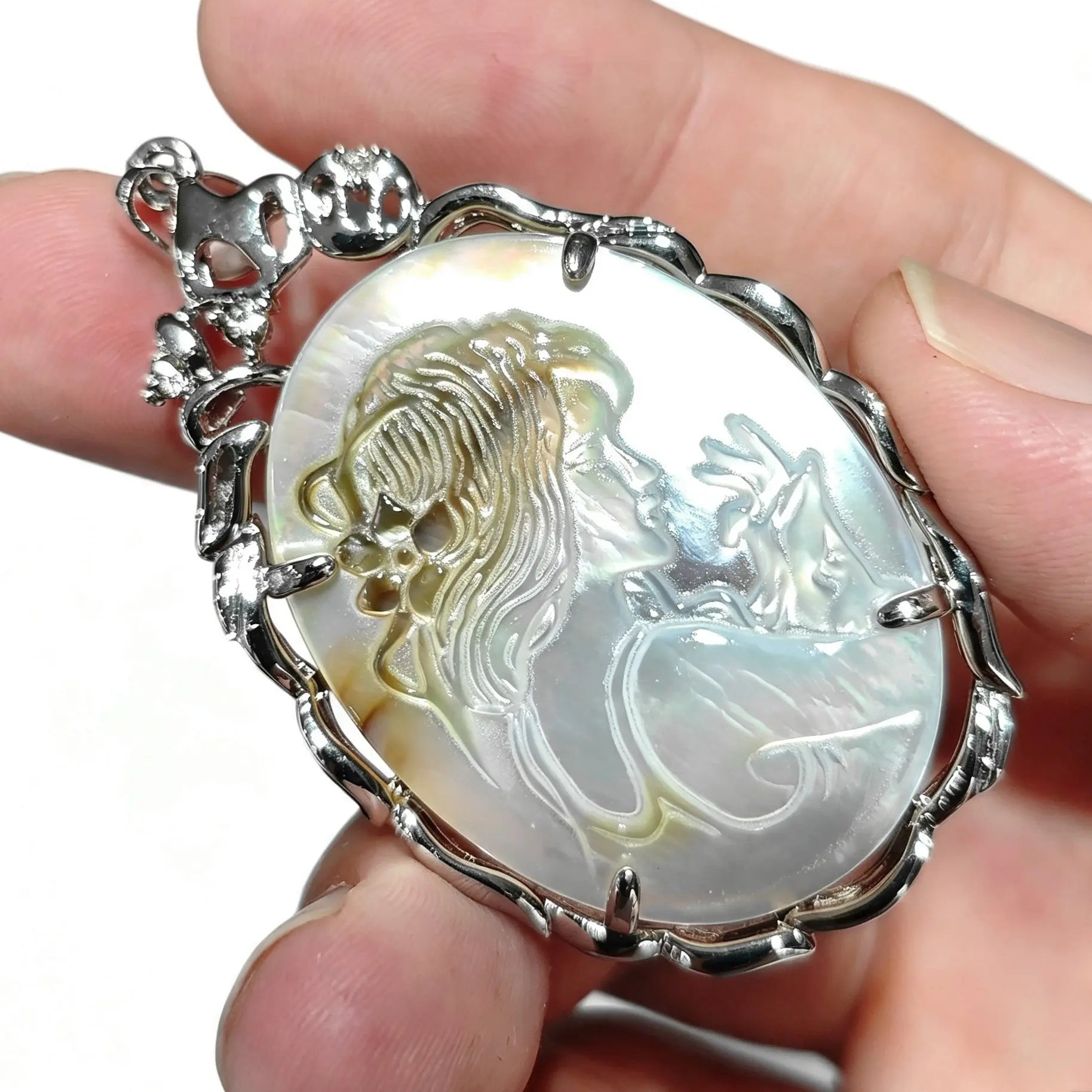 Close up view of carved mother of pearl cameo pendant held between fingers displaying ornate silver tone prongs