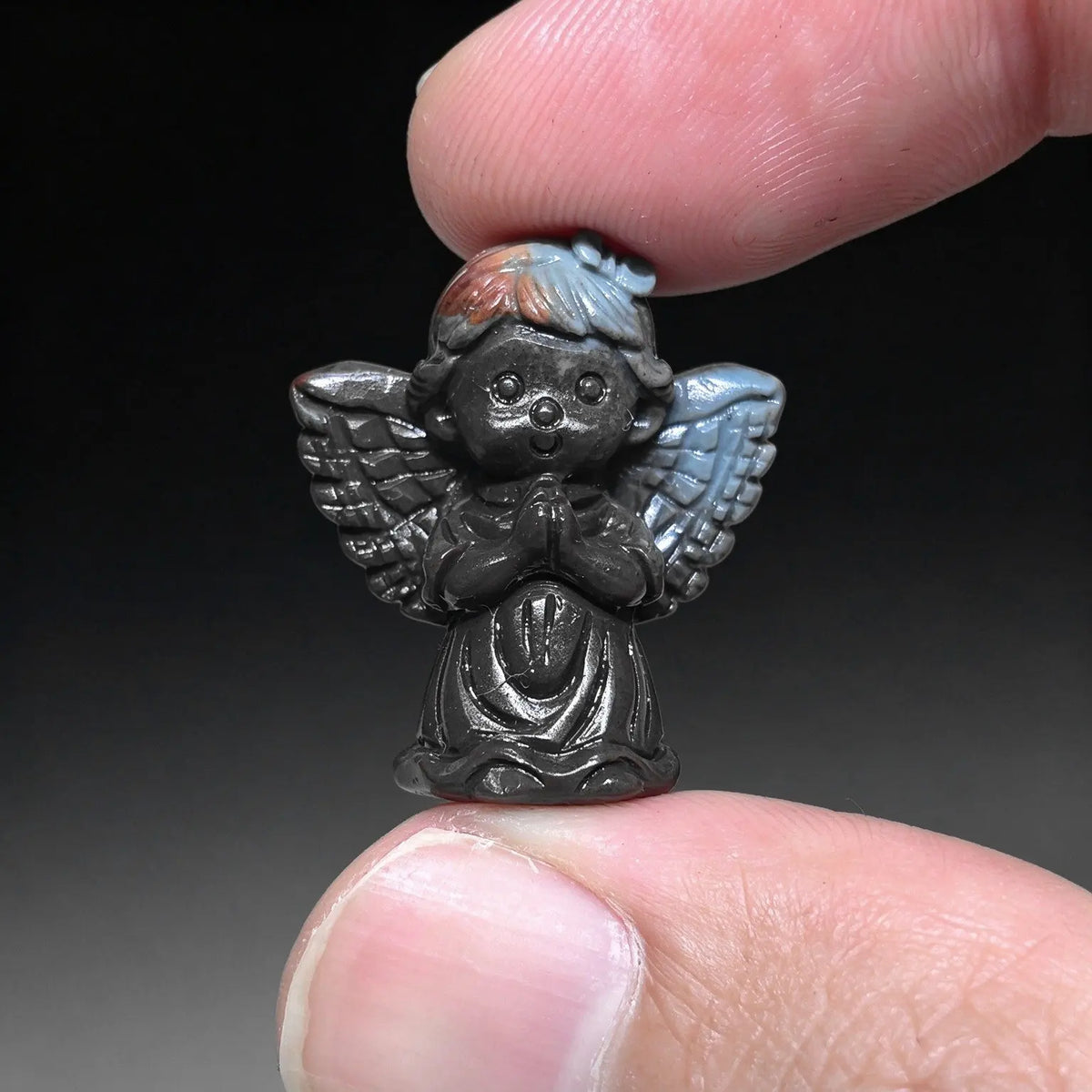 Front view of a tiny praying angel figurine in dark charcoal and slate gray held between two fingertips