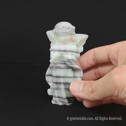 Hand holding an approx 10cm zebra stone praying angel carving to showcase its actual physical size, matte finish, and monochrome pattern for wholesale buyers