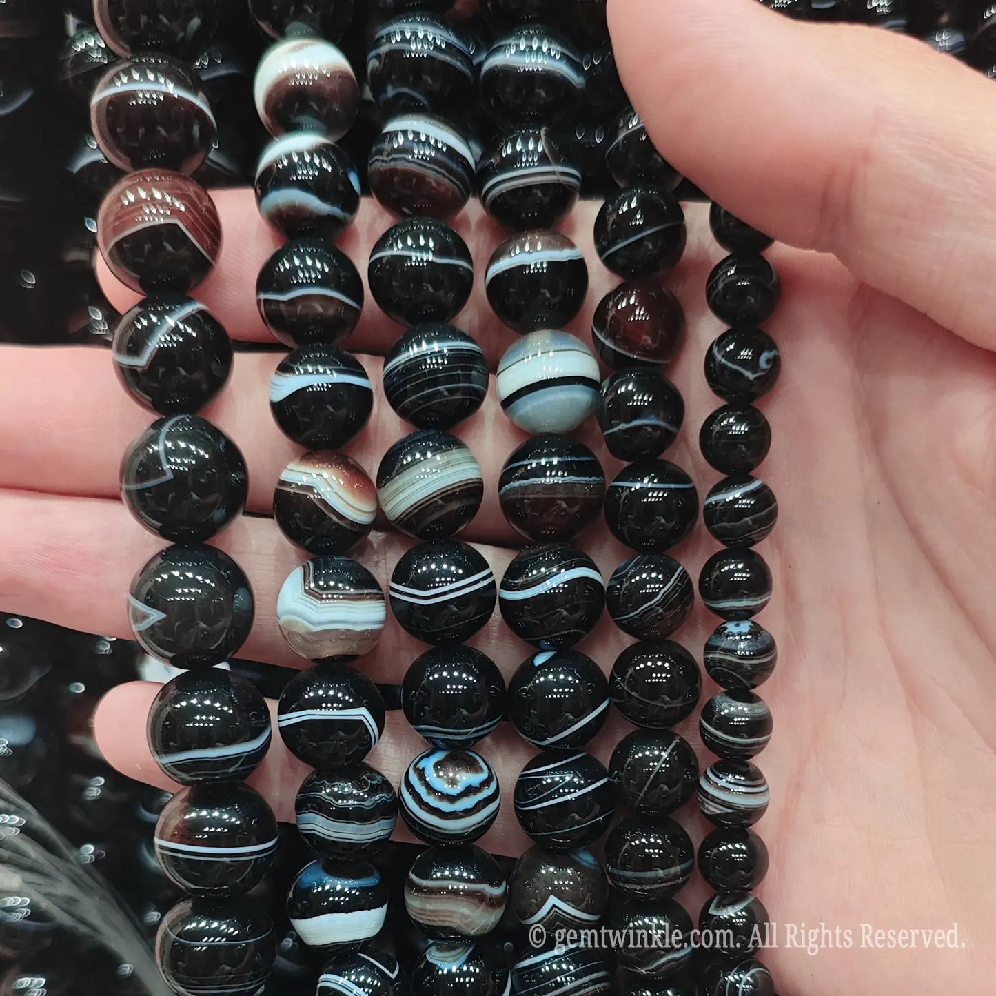 Hand holding natural dark brown banded agate loose beads strands under bright light highlighting genuine silk stripes and polished finish
