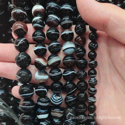 Hand holding natural dark brown banded agate loose beads strands under bright light highlighting genuine silk stripes and polished finish