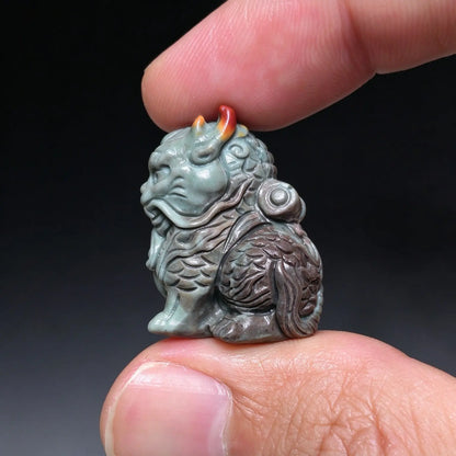 Side profile view of a carved Kirin beast figurine in slate gray stone with red horn accents held between fingertips