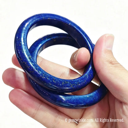 Angled hand view of two solid lapis lazuli crystal bangles showcasing vibrant ultramarine blue and smooth inner contour