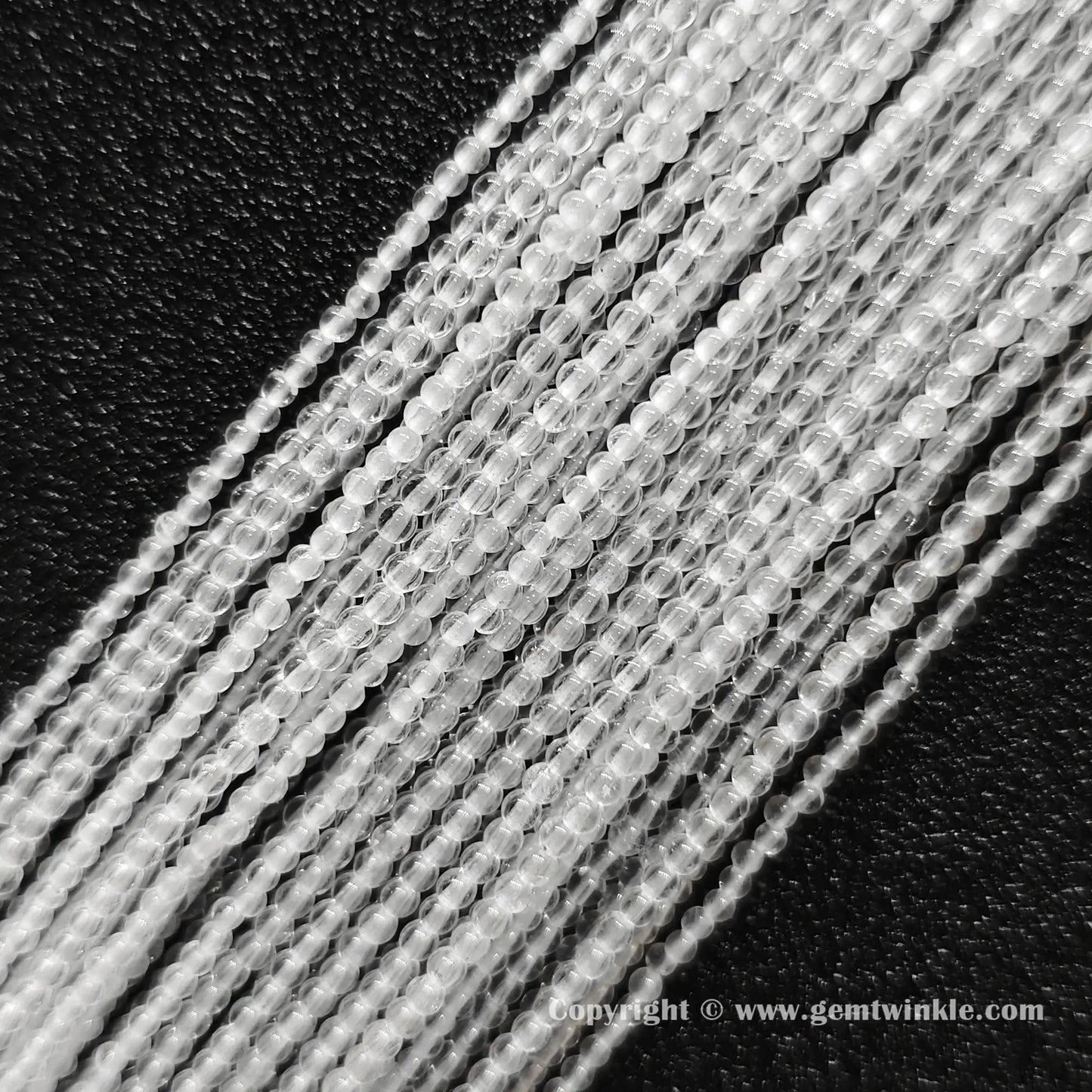 Angled view of 2mm and 3mm micro rock crystal round bead strands laid parallel on dark surface