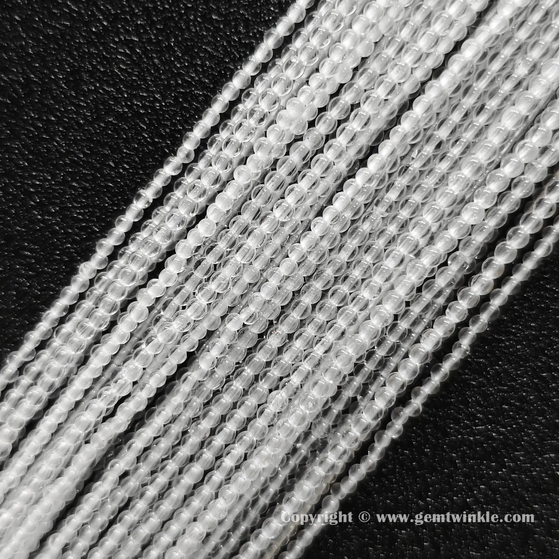 Angled view of 2mm and 3mm micro rock crystal round bead strands laid parallel on dark surface