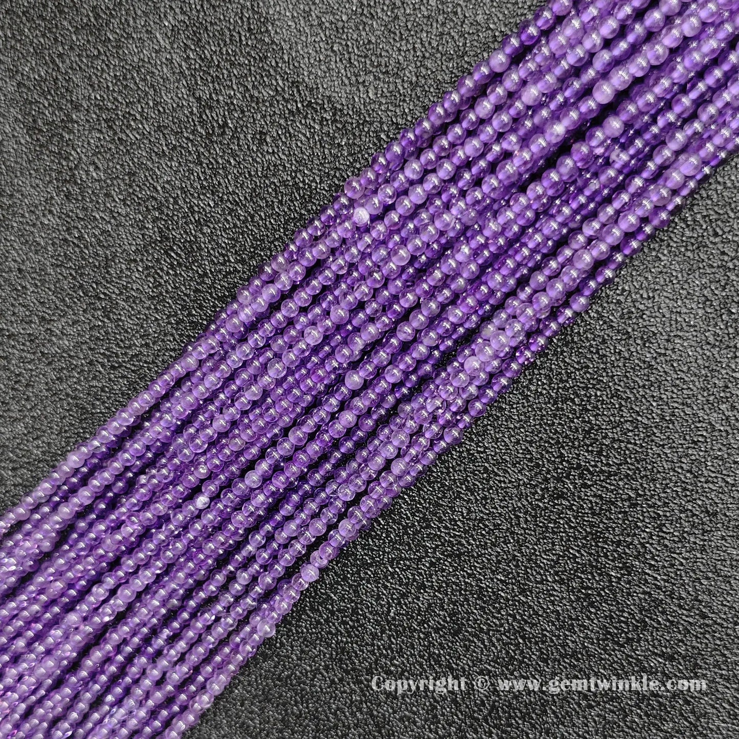 Angled view of 2mm mini amethyst gemstone strands laid parallel on dark textured surface