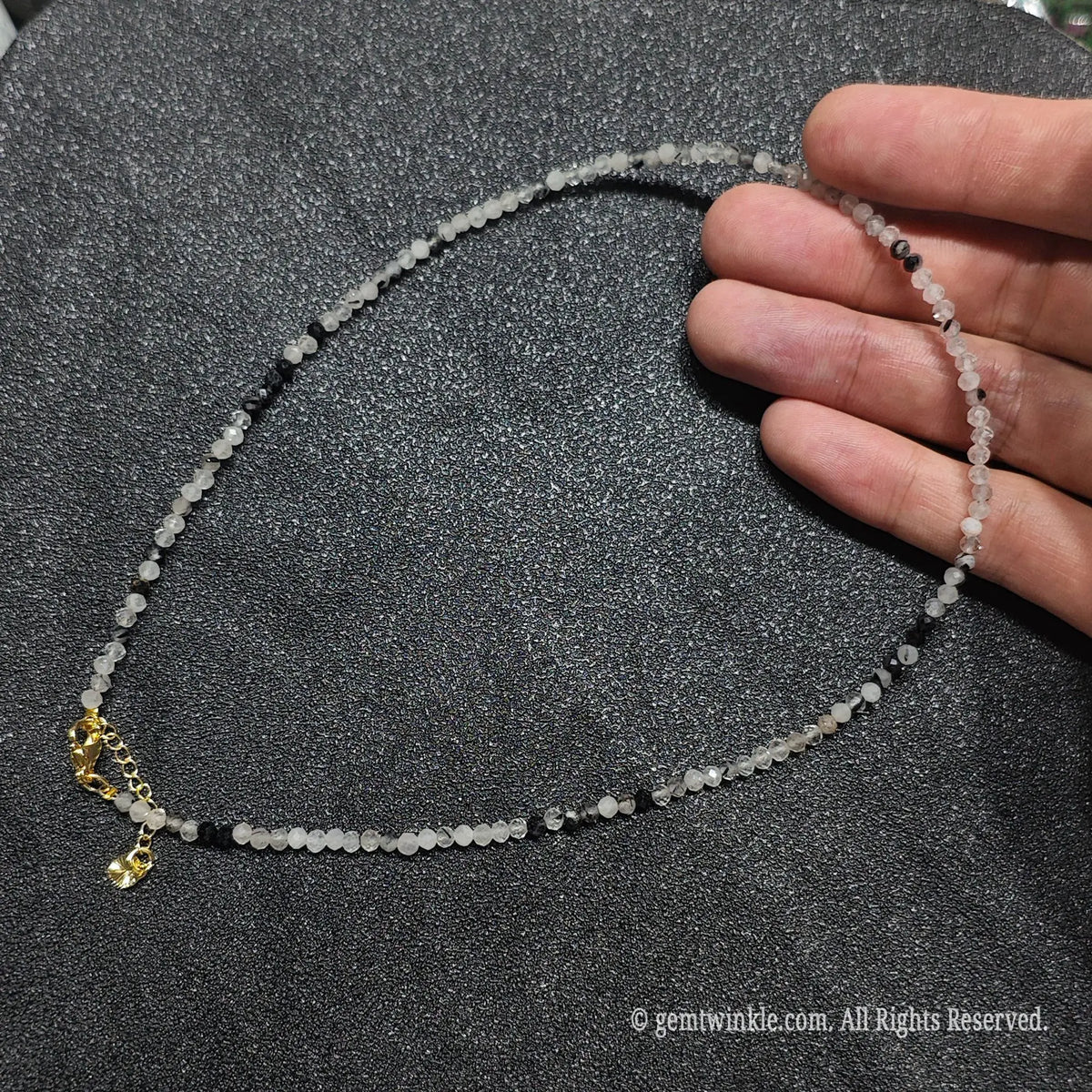 Hand holding 3mm micro faceted black rutilated quartz necklace strand showing misty grey quartz with black needles