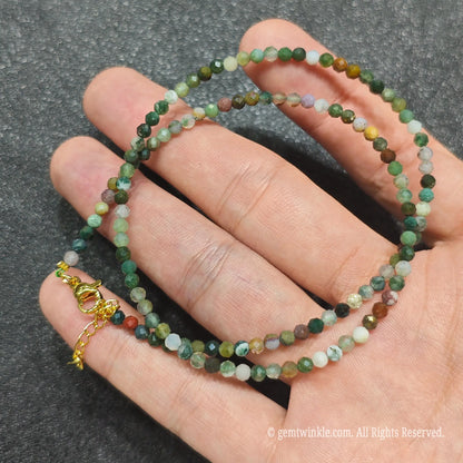 A beaded necklace made of small faceted gemstone beads in earthy green, brown, gray, and white tones, with a gold clasp, draped over a person's hand against a dark textured background.