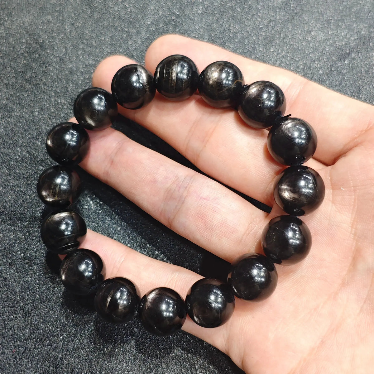Close up view of a hand holding a natural hypersthene bracelet featuring 12mm dark beads with silver golden schiller sheen
