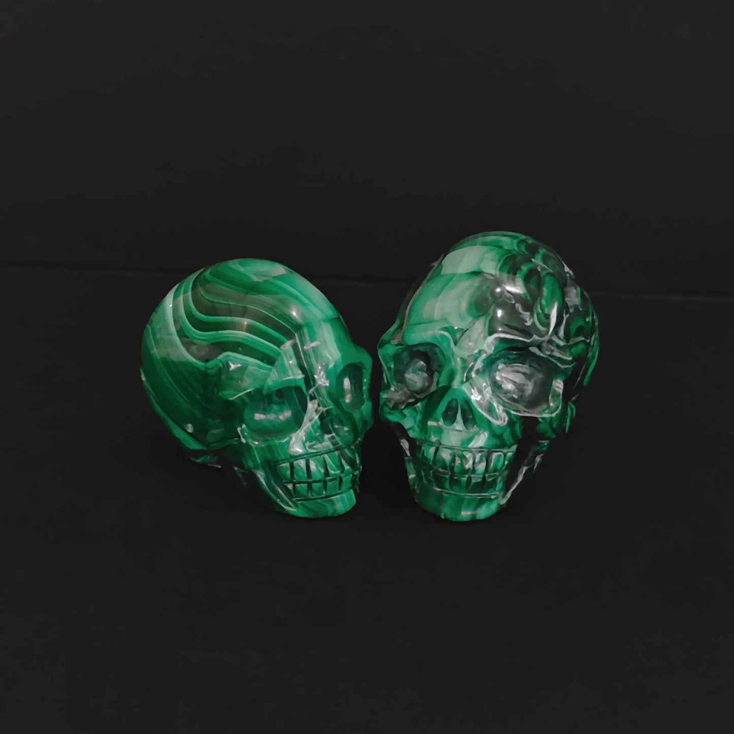 Pair of natural green malachite crystal skull carvings with vivid banded patterns sitting on a black background