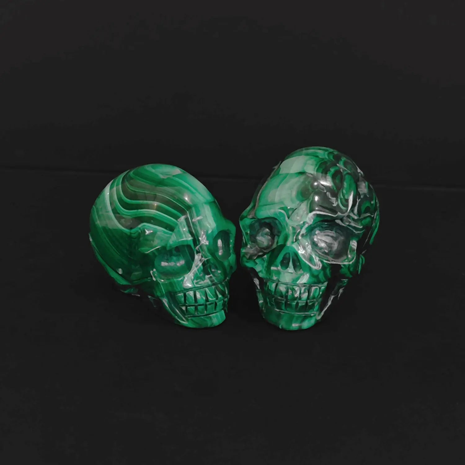 Pair of natural green malachite crystal skull carvings with vivid banded patterns sitting on a black background