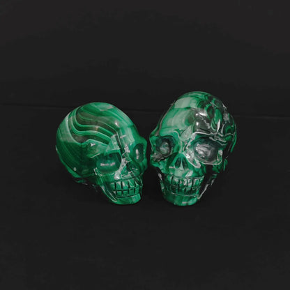 Pair of natural green malachite crystal skull carvings with vivid banded patterns sitting on a black background