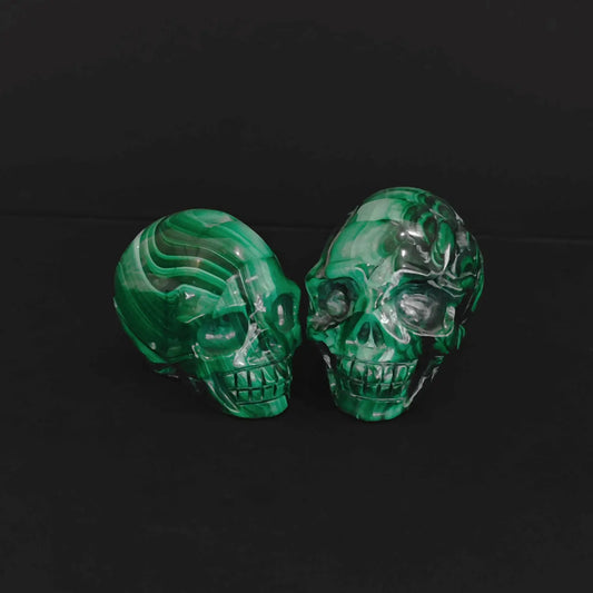 Pair of natural green malachite crystal skull carvings with vivid banded patterns sitting on a black background