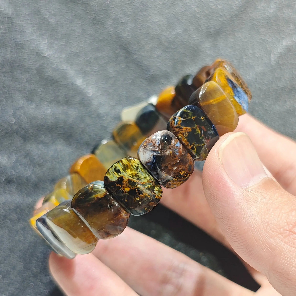 Hand holding a natural multicolor pietersite unisex boho bracelet featuring rectangle beads with golden blue and brown swirling patterns[cite: 6]