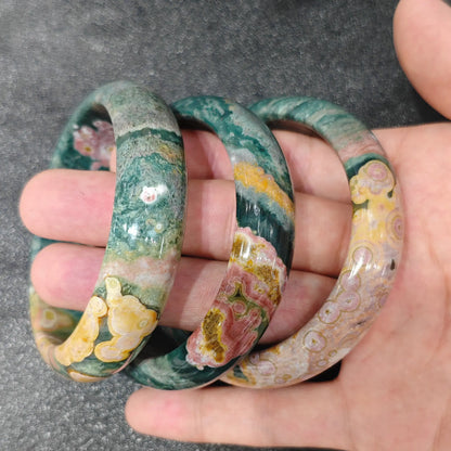 Hand holding three natural ocean jasper bangles showing dark green, mustard yellow, and pink orbicular patterns