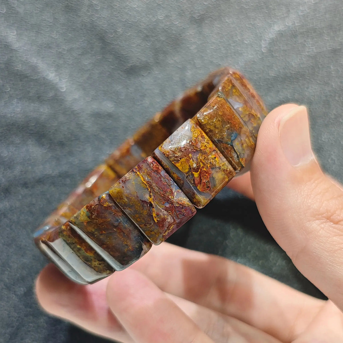 Hand holding a natural red fiery pietersite unisex dark boho plate bead bracelet with amber and golden swirls