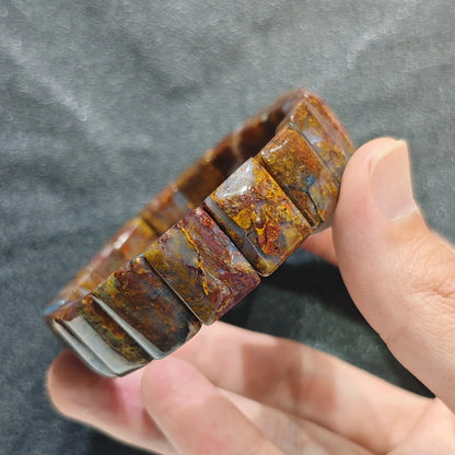 Hand holding a natural red fiery pietersite unisex dark boho plate bead bracelet with amber and golden swirls