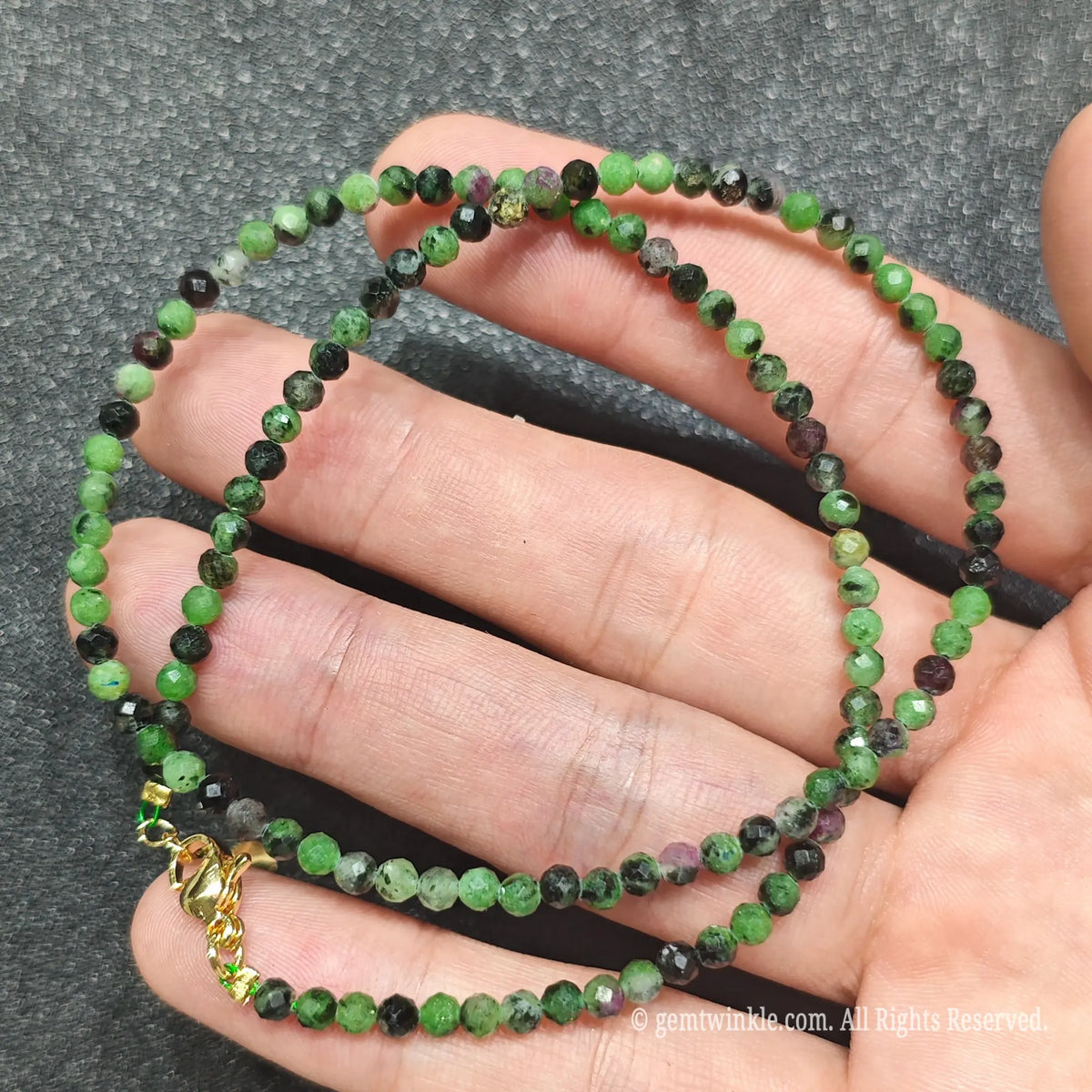 A beaded necklace made of small faceted green gemstone beads with some black and purple marbled accents, held in a person's hand. The necklace has a gold clasp.