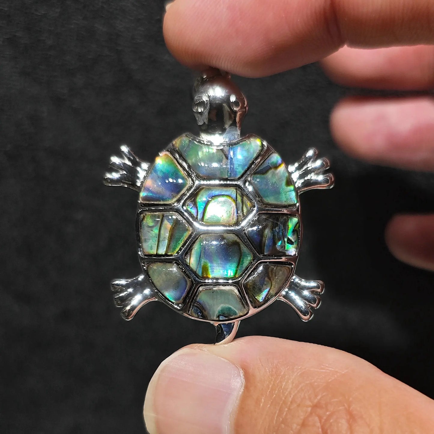 Fingers holding a 35mm sea turtle pendant with colorful iridescent abalone shell mosaic inlay framed in silver tone alloy metal