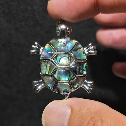 Fingers holding a 35mm sea turtle pendant with colorful iridescent abalone shell mosaic inlay framed in silver tone alloy metal