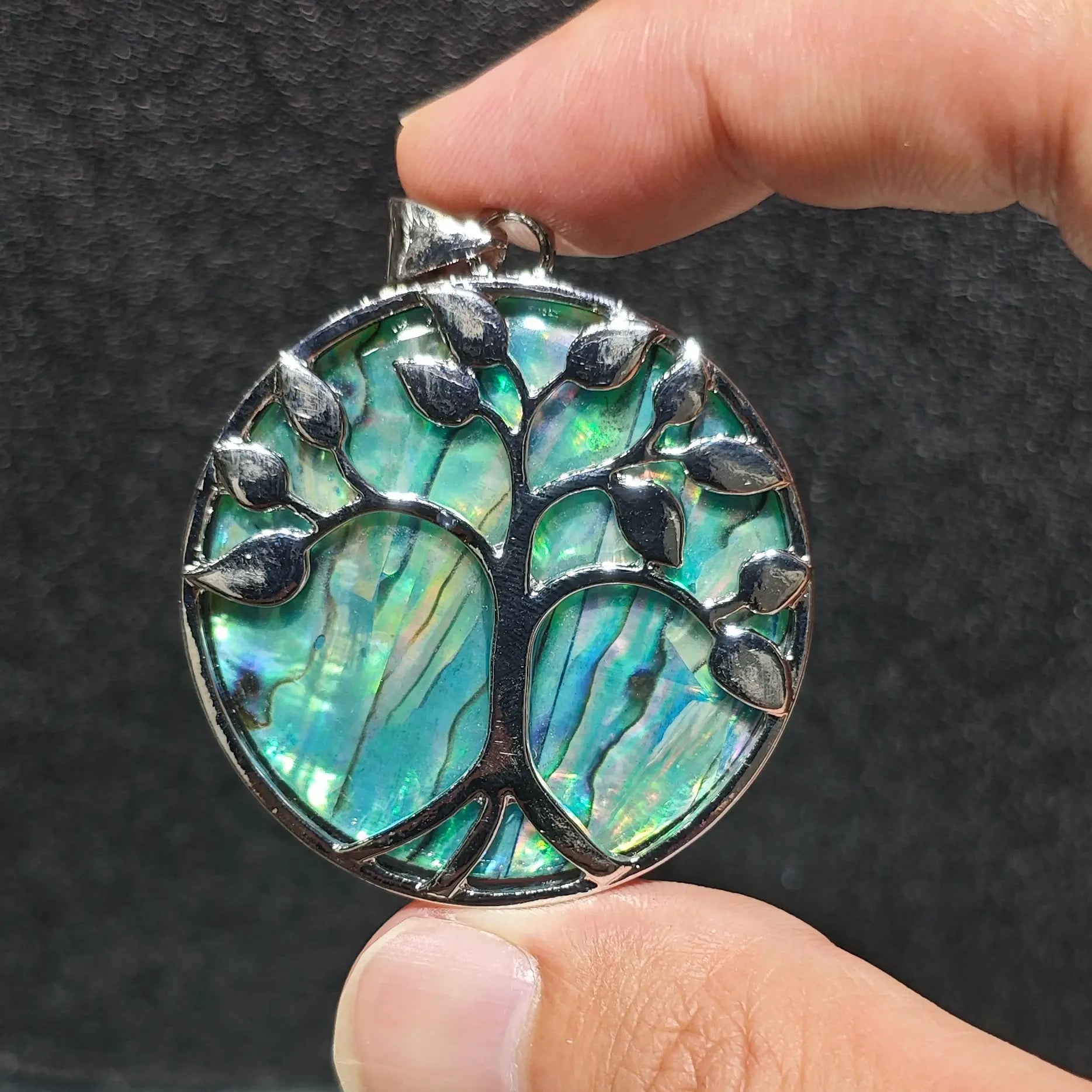 Fingers holding a 5cm round Tree of Life pendant with vibrant blue abalone shell inlay set under silver tone alloy branches