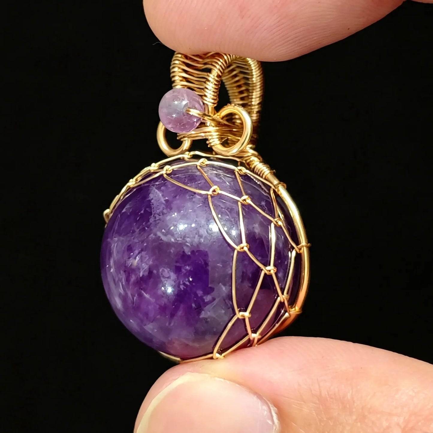 Fingers holding a 20mm natural amethyst sphere pendant enclosed in gold wire wrapping with top accent bead