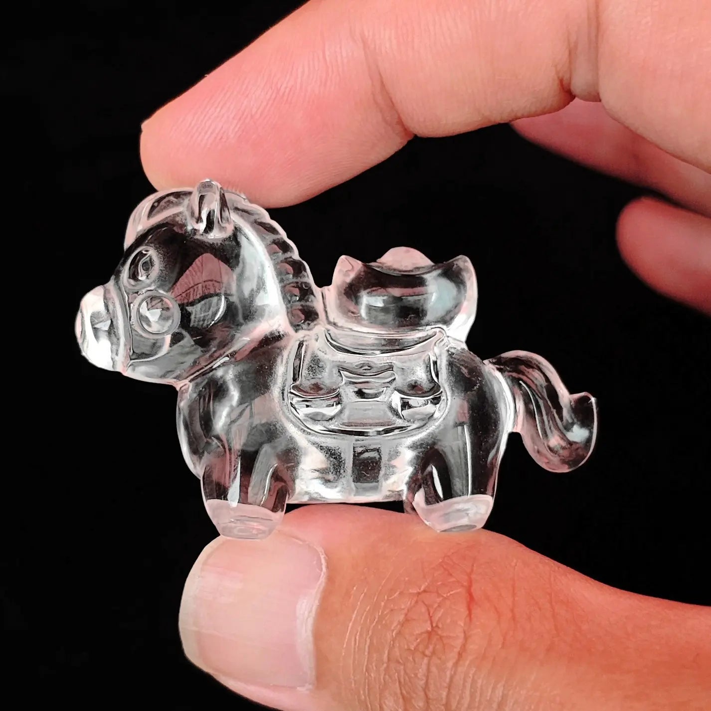 Fingers holding a high clarity precision carved natural clear quartz horse carrying a wealth ingot side view against a dark background