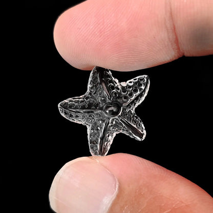 Fingers holding a high clarity precision carved natural clear quartz starfish gemstone with textured relief against a black background