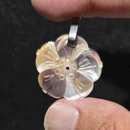 Fingers holding a 25mm precision carved natural grey mother of pearl plum blossom pendant displaying silvery shell luster against a dark background