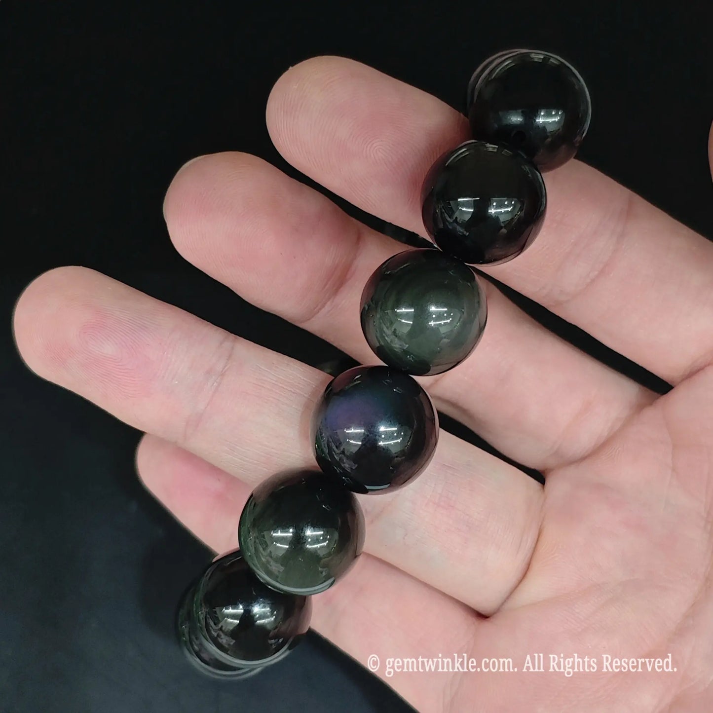 Natural obsidian beads string held under studio lighting displaying iridescent purple and green sheen eyes