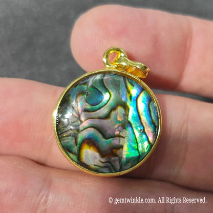 Close up view of single sided 2cm round abalone shell charm with gold tone rim displaying blue and green peacock reflections