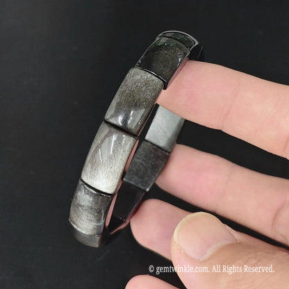 Angle view of natural silver obsidian rectangular plate bead stretch bracelet held showing silver flash reflections