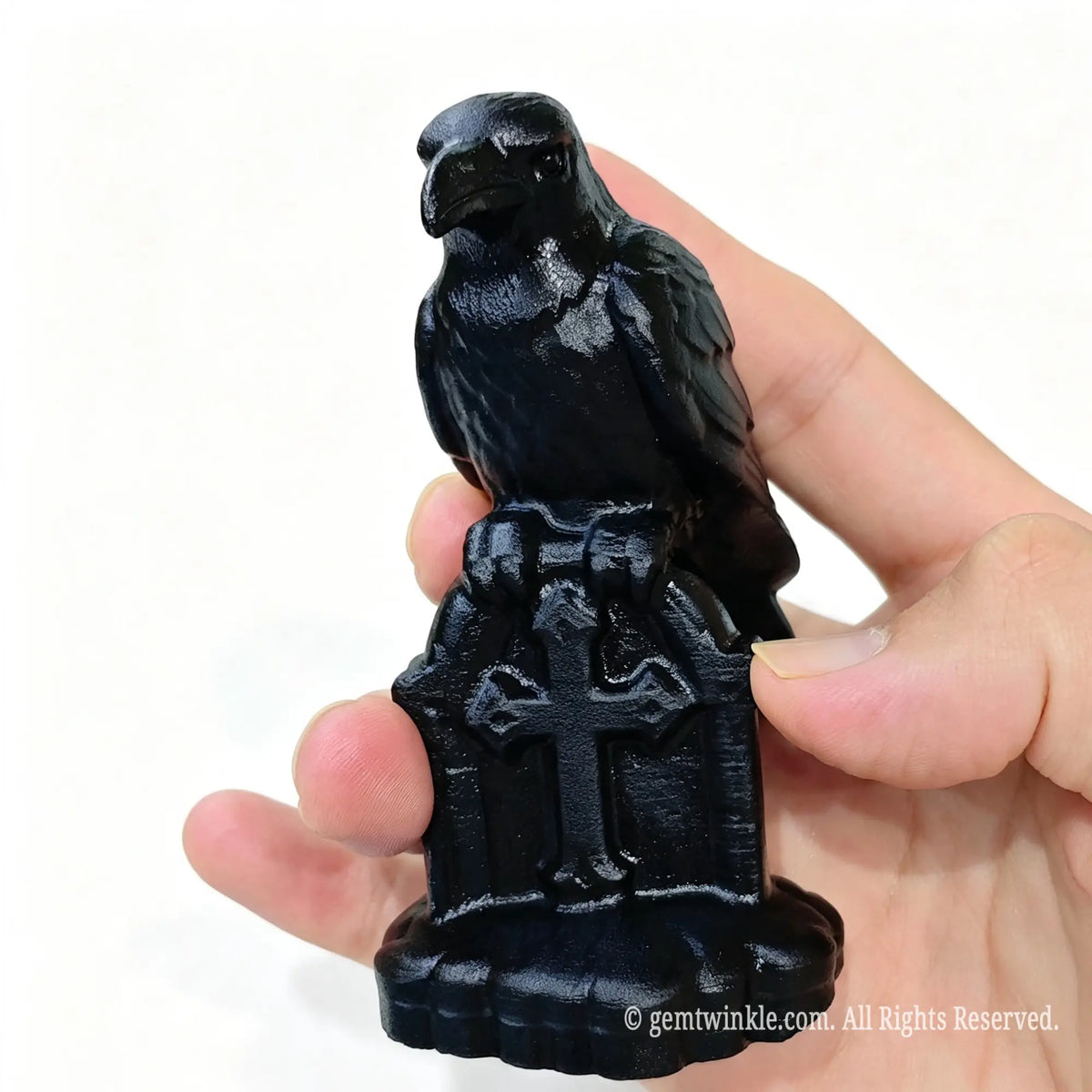 Frontal view of black obsidian raven sculpture perched on carved cross tombstone base held in hand
