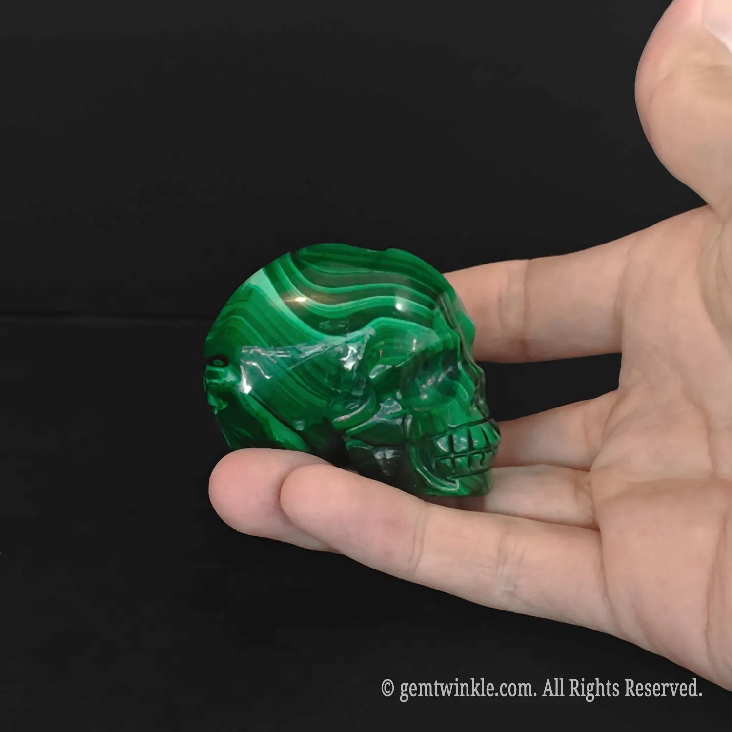Polished malachite mineral skull carving held in a hand highlighting dark green eye sockets and distinctive swirls
