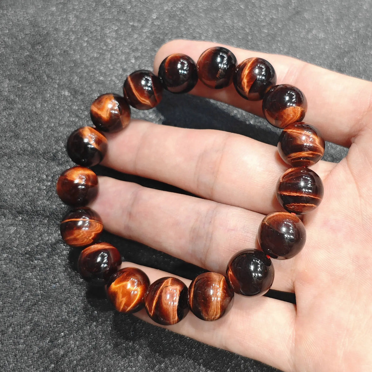 Close up view of a hand holding a premium red tiger's eye bracelet with vibrant 12mm beads and lightning flash chatoyancy